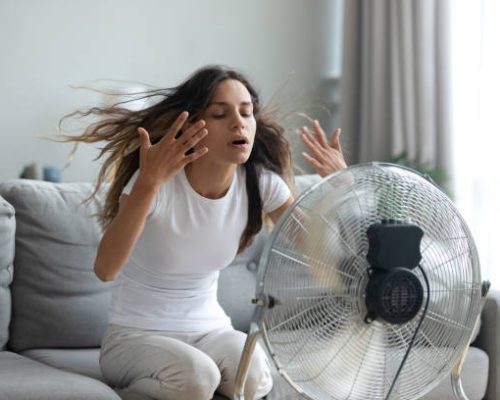 In living room without air-conditioner tired from summer heat young woman turned on floor ventilator waving her hands to cool herself, female sitting on couch suffers from unbearable too hot weather